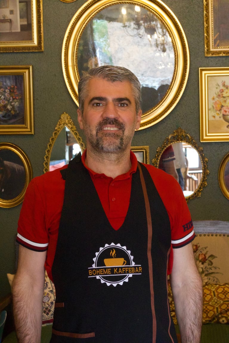 Man wearing red and black apron with Boheme Kaffebar logo, standing in front of ornate gold-framed mirrors and artwork