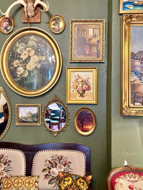 Gallery wall with framed artwork and decorative objects on sage green wall, featuring floral paintings, ornate gold frames, and classical decor above upholstered furniture
