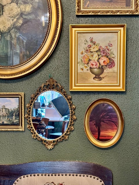 Gallery wall with ornate framed artwork including paintings, mirrors, and landscape prints arranged on sage green wall above upholstered furniture