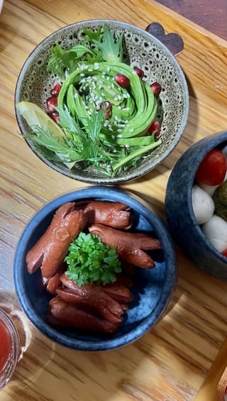 Two bowls of food on wooden surface: green avocado paste with pomegranate seeds and sesame in gray speckled bowl, red sausages with parsley garnish in blue bowl