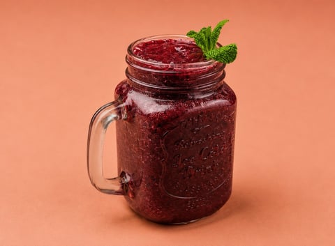 A glass mason jar filled with deep red berry smoothie or juice, garnished with fresh mint leaves on top, against an orange background.
