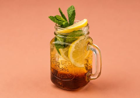 Mason jar filled with cola and ice, topped with fresh mint sprigs and a lemon slice on a coral background