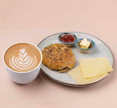 Cappuccino with latte art beside a plate with chocolate chip scone, cheese slice, jam, and butter on beige background