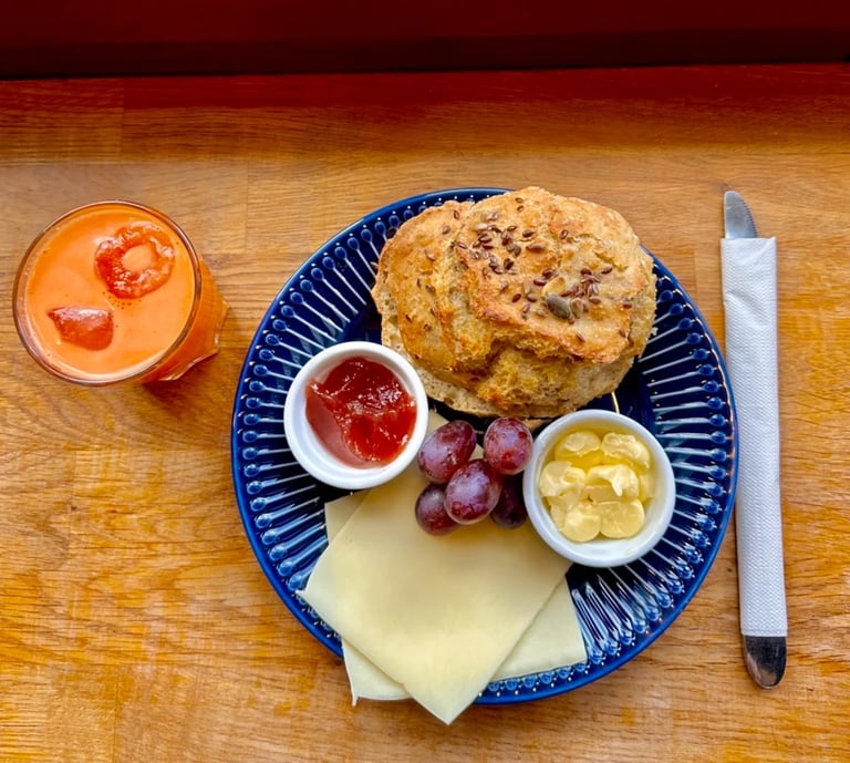 Breakfast plate with scone, cheese, red grapes, jam, butter, and a gazpacho soup on wooden table