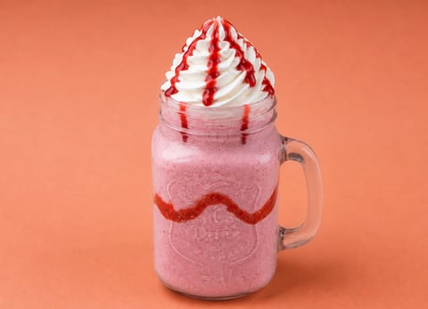 Pink milkshake in a glass jar topped with whipped cream and red syrup drizzle on orange background