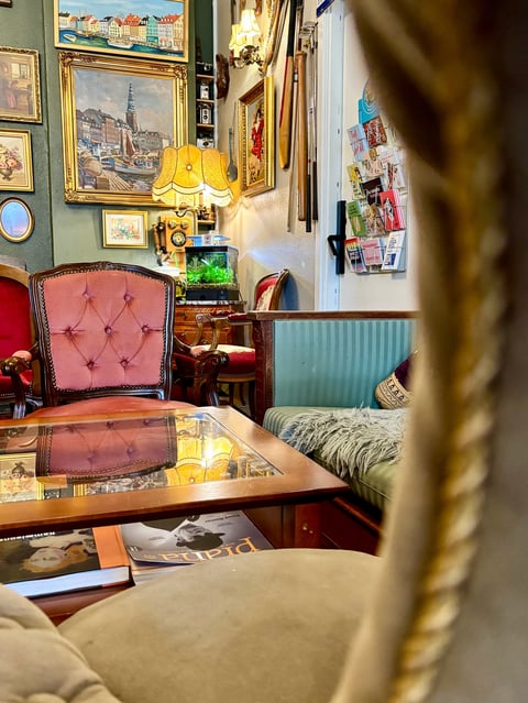 Eclectic interior room with pink velvet chair, wooden desk, framed artwork, yellow lamp, and vintage decorative items on walls and surfaces